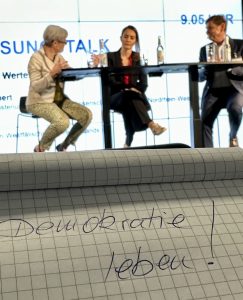 Podium mit Diskutanten. Davor ein Schreibblock mit der handschriftlichen Notiz: Demokratie leben!