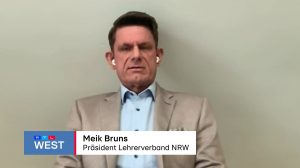 Zoff um die Bildungspolitik im Westen: Ein Mann mit hellem Sakko und Kopfhörern schaut in eine Kamera. In einer Bauchbinde steht sein Name und seine Funktion.
